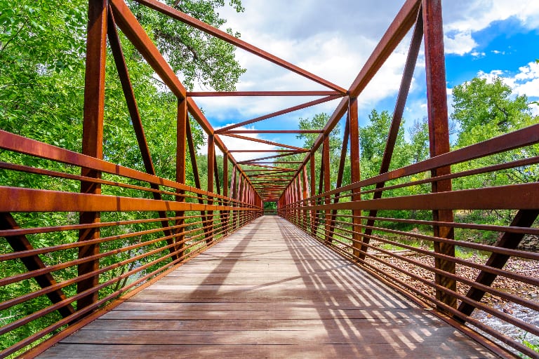 A pedestrian bridge over the Cache la Poudre River in Fort Collins, framed by lush trees under a bright sky