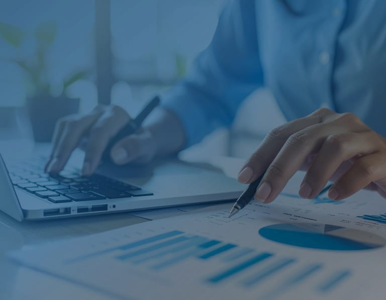Hands working at a laptop with printed reports, a pen, and forms on the desk