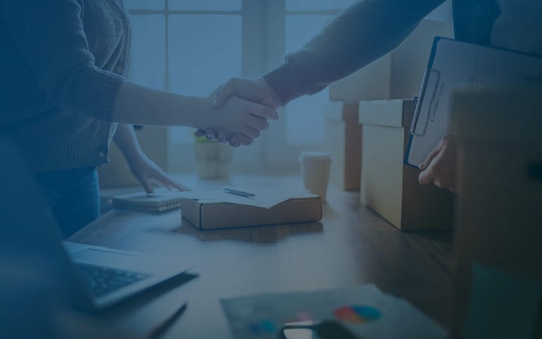 A handshake over a desk with a laptop, paperwork, and packing boxes — a small business partnership in motion